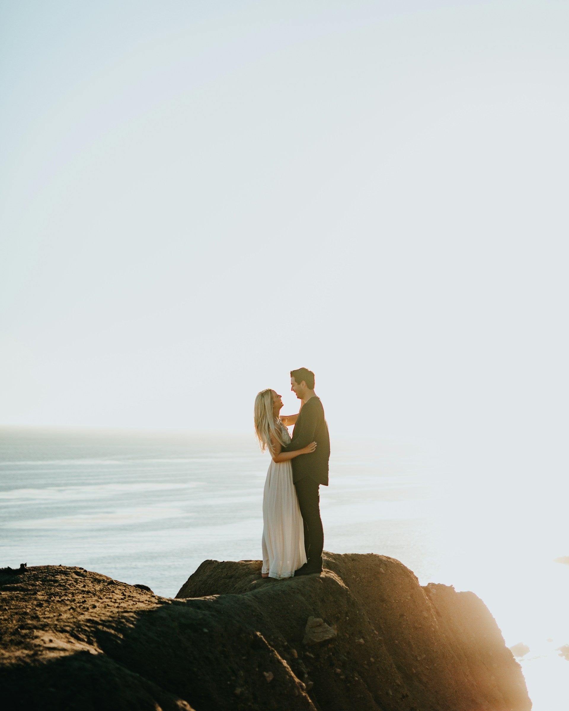 Couple embracing on a rocky outcrop by the ocean with a clear sky.